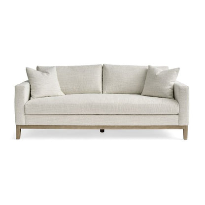 The Capri Sofa is a minimalist design featuring a beige finish, two seat cushions, and four matching throw pillows, all supported by short wooden legs. It is showcased against a plain white background.