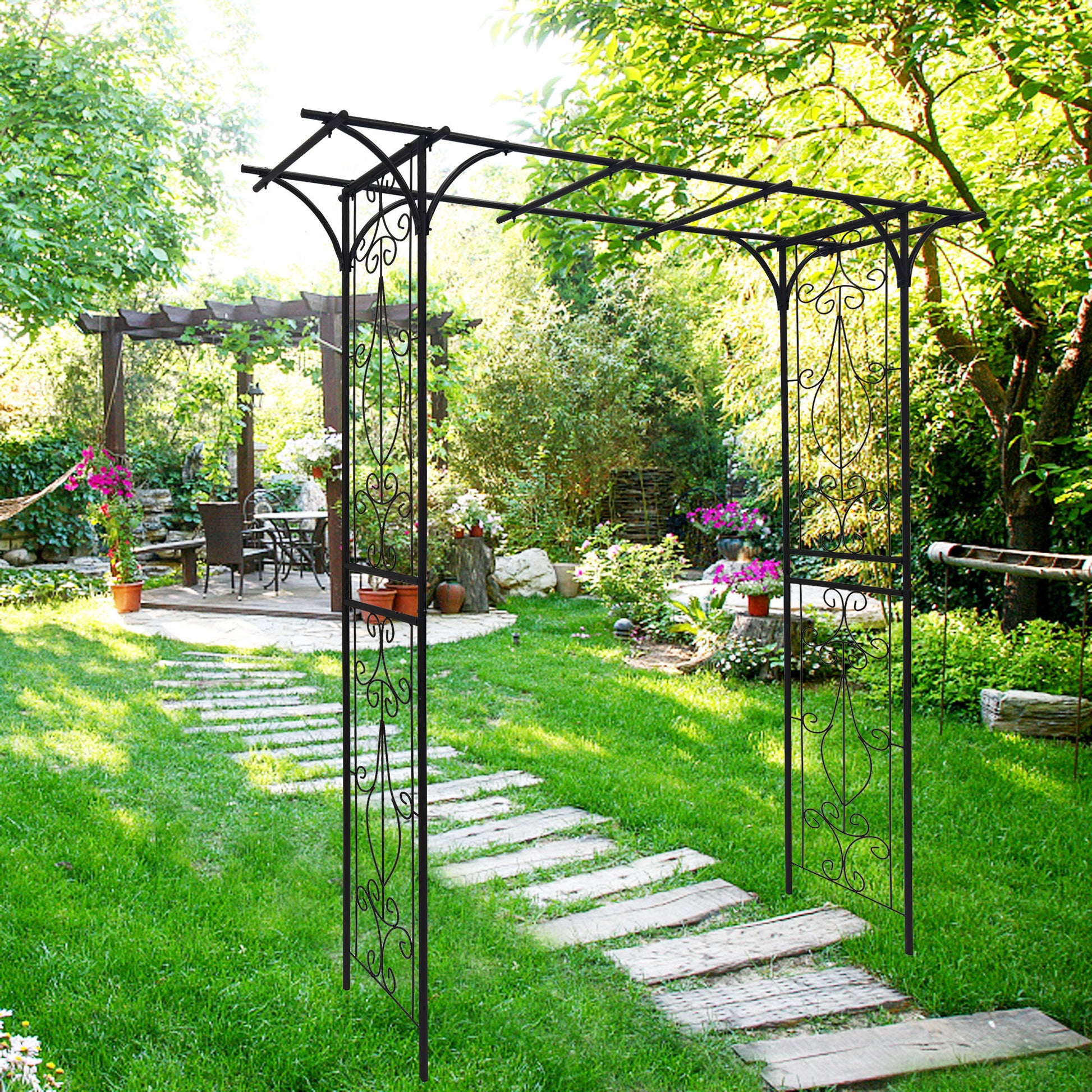 The Metal Garden Arch, designed for climbing plants, elegantly frames a stone path to a shaded seating area. The lush garden features green foliage, flowers, and a wooden pergola while sunlight filters through the trees, creating dappled light on the pathway.