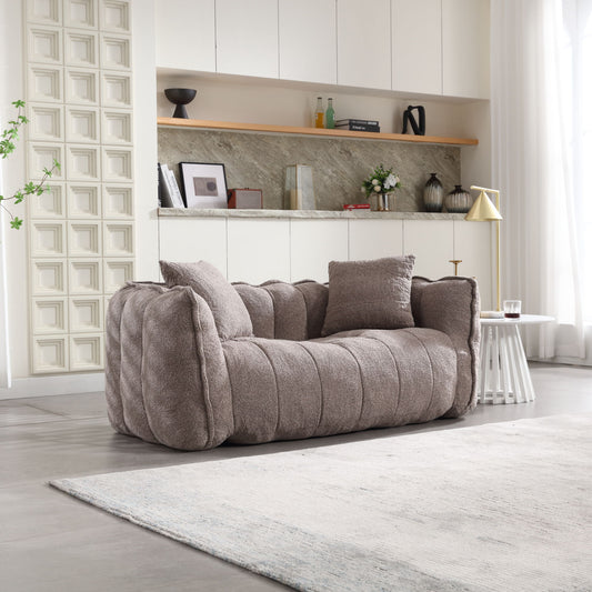 A soft, gray beanbag chair with high resilience foam sits on a light rug in a modern living room. In the background are white cabinets, a marble-look backsplash, and shelves holding decor like a small lamp, books, and plants on the side table.