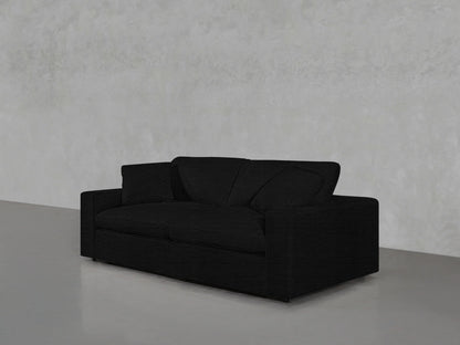 A black 2-Seat Modular Loveseat with a modern design sits on a polished concrete floor against a light gray textured wall. The loveseat features simple lines and two large pillows.