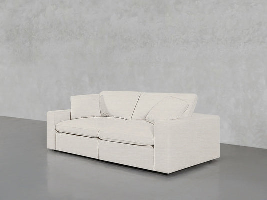 A 2-Seat Modular Loveseat in a modern, light gray finish, featuring two seat cushions and two back cushions, sits against a textured gray wall on a smooth, dark gray floor. The design is minimalist, showcasing a low-profile and rectangular shape.