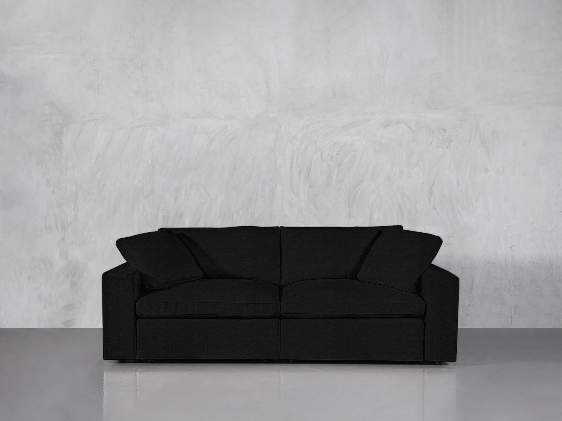 A black 2-Seat Modular Loveseat with two cushions is placed against a light gray concrete wall on a shiny gray floor. The loveseat features a modern and minimalist design.