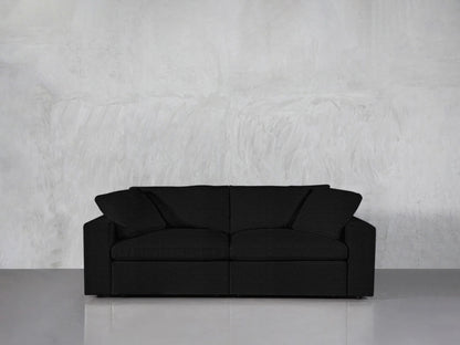 A black 2-Seat Modular Loveseat with two cushions is placed against a light gray concrete wall on a shiny gray floor. The loveseat features a modern and minimalist design.