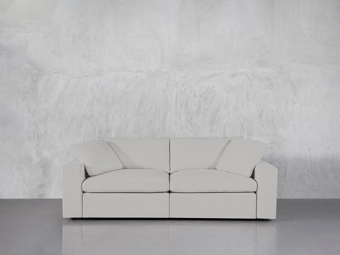 A modern 2-Seat Modular Loveseat in light gray, featuring three cushions, is positioned against a textured concrete wall on a smooth gray floor, conveying a minimalist and simple aesthetic.