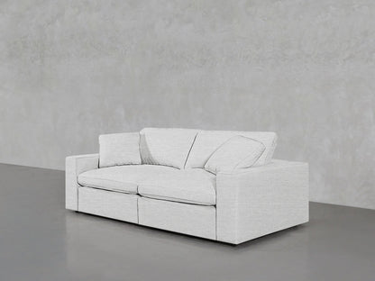 A sleek, white 2-Seat Modular Loveseat with cushions is set against a plain gray textured wall. The floor is smooth and gray, enhancing the minimalist aesthetic.