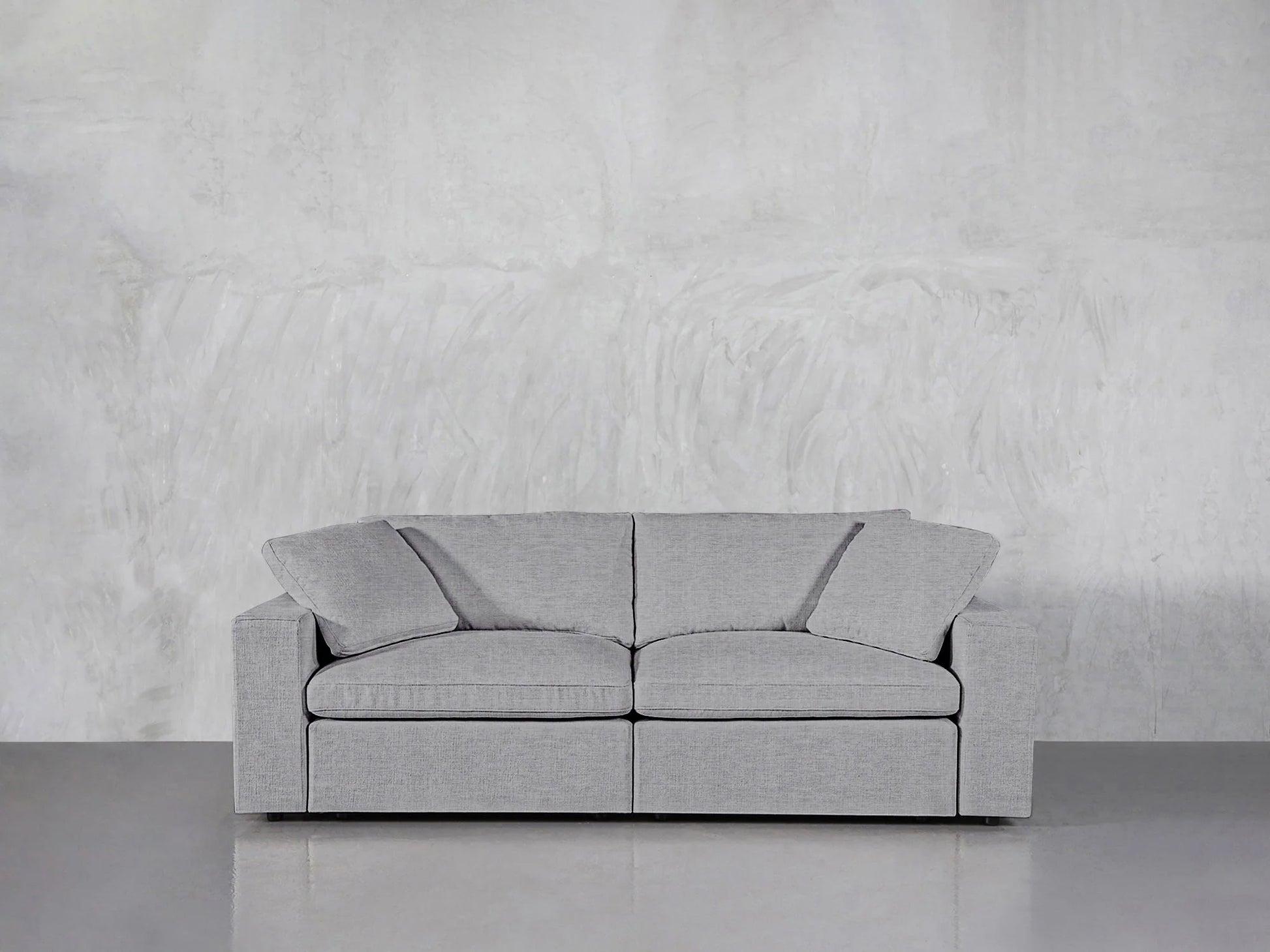 The 2-Seat Modular Loveseat, featuring a modern gray design with clean lines and plush cushions, is placed in a minimalist room with a light gray textured wall and sleek concrete floor, creating a simple and contemporary setting.