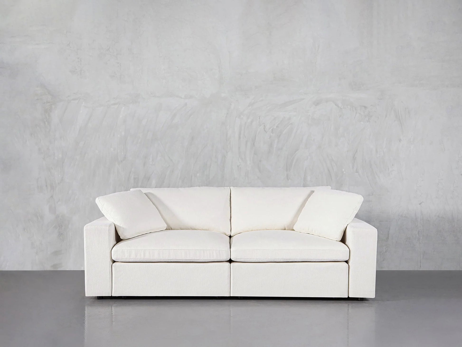 A 2-Seat Modular Loveseat, characterized by its modern white finish and clean lines, is placed against a plain gray wall. This sleek, minimalist design features two seat cushions and a high backrest, adding a contemporary look to the setting.