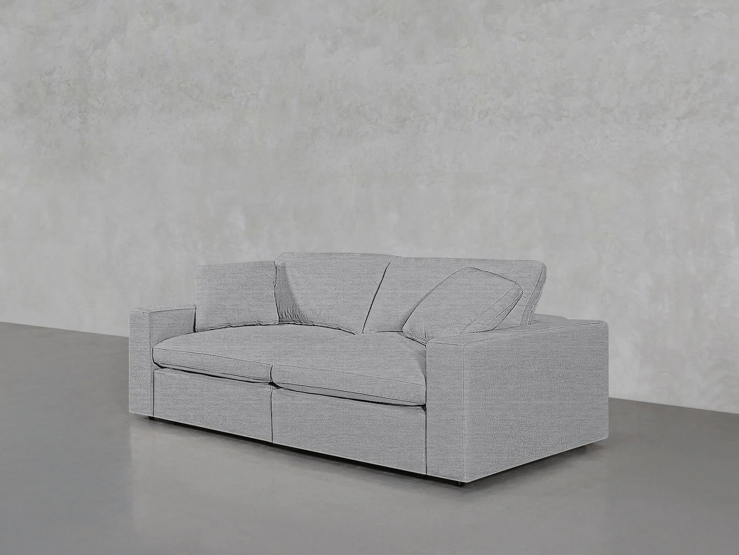 A 2-Seat Modular Loveseat with a minimalist design and two cushions sits on a smooth concrete floor against a light grey textured wall. This loveseat boasts clean lines and a modern aesthetic, perfect for contemporary spaces.