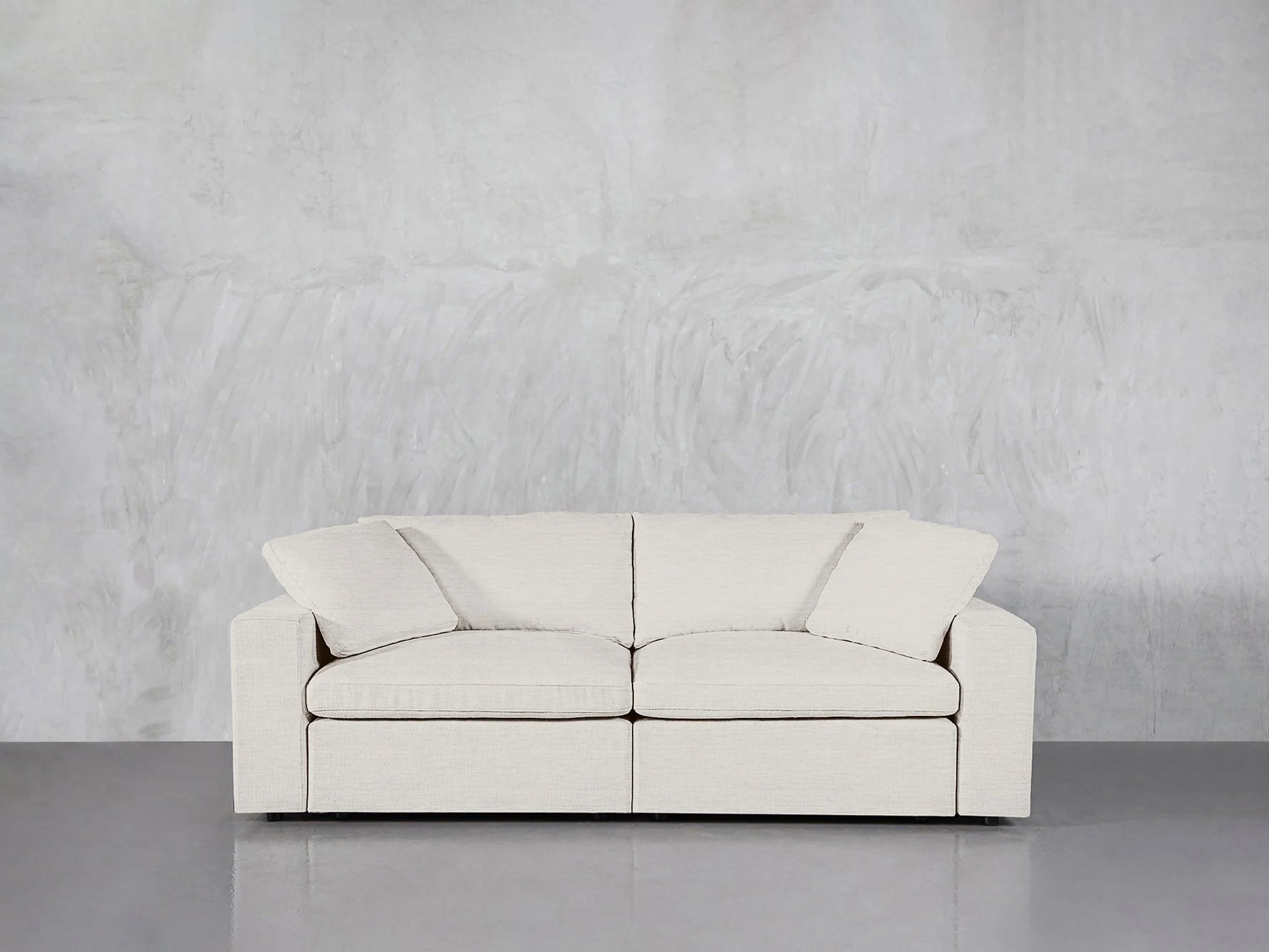 A modern, white 2-Seat Modular Loveseat with seat and back cushions is set against a plain gray, textured wall. The smooth grey floor enhances the minimalist and clean aesthetic.