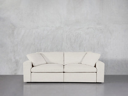 A modern, white 2-Seat Modular Loveseat with seat and back cushions is set against a plain gray, textured wall. The smooth grey floor enhances the minimalist and clean aesthetic.