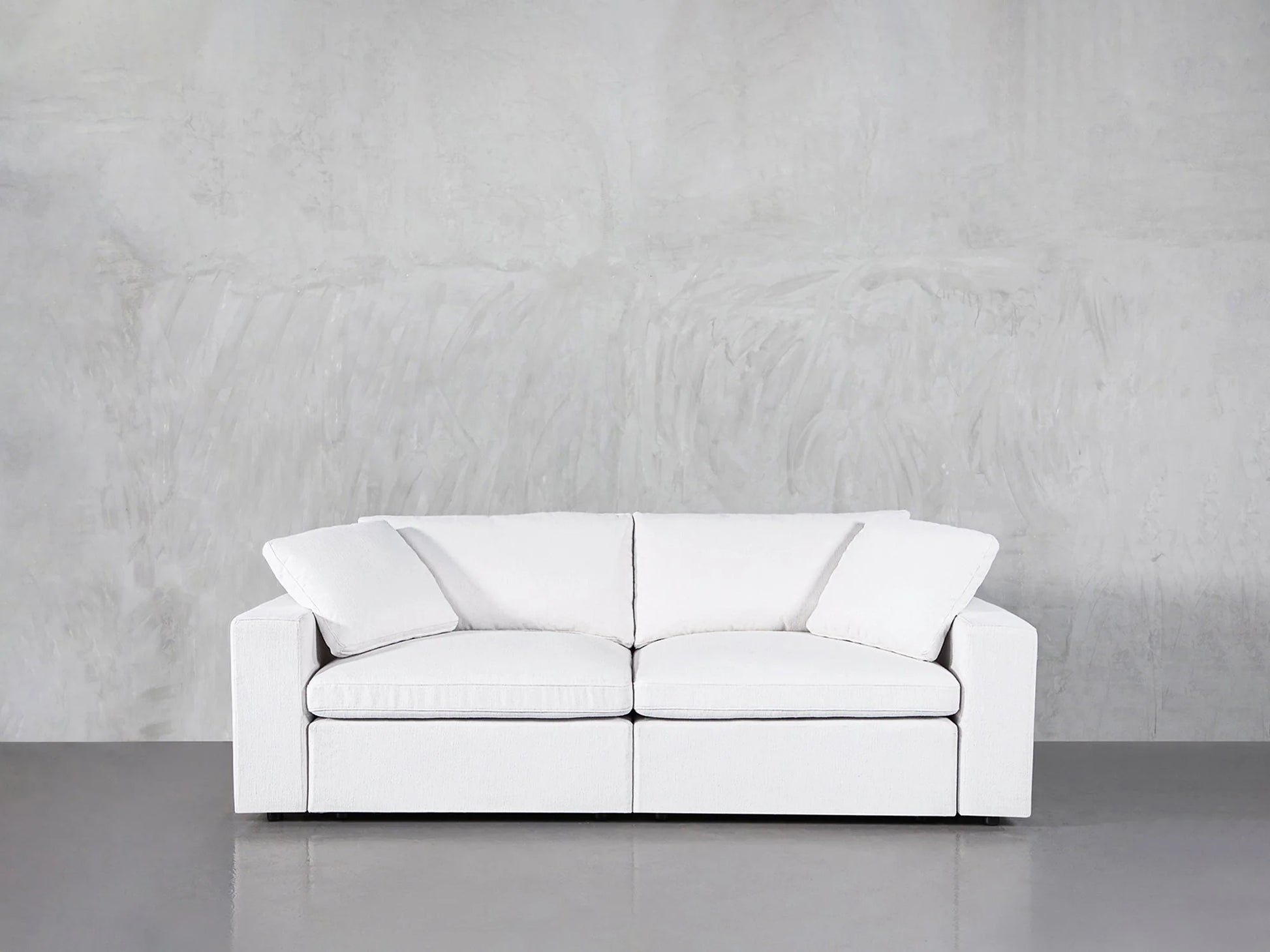 A white 2-seat modular loveseat in a modern minimalist setting against a textured gray wall. It features two seat cushions and two large back cushions, offering a clean and simple design.