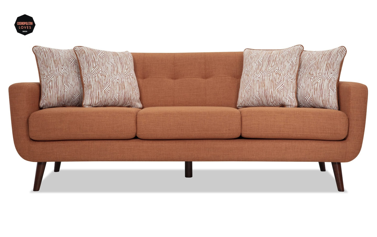 The Bettie 86'' Sofa is a modern piece featuring an orange-brown color with three seat cushions and patterned throw pillows. It boasts wooden legs and a tufted backrest, offering a stylish and comfortable appearance.