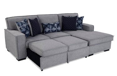 The Playscape Gray 94'' 2 Piece Pop-Up Sleeper Chaise Sectional is a modern piece featuring clean lines and a minimalist design, complete with a chaise lounge. It is arranged neatly with several decorative cushions, including dark blue and patterned pillows.