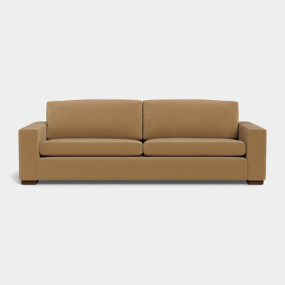 The Barton Sofa, a modern piece with a boxy design, is made from brown leather and showcases two seat cushions and two back cushions made from the same material. It features wide armrests and dark wooden feet, all displayed against a plain white background.