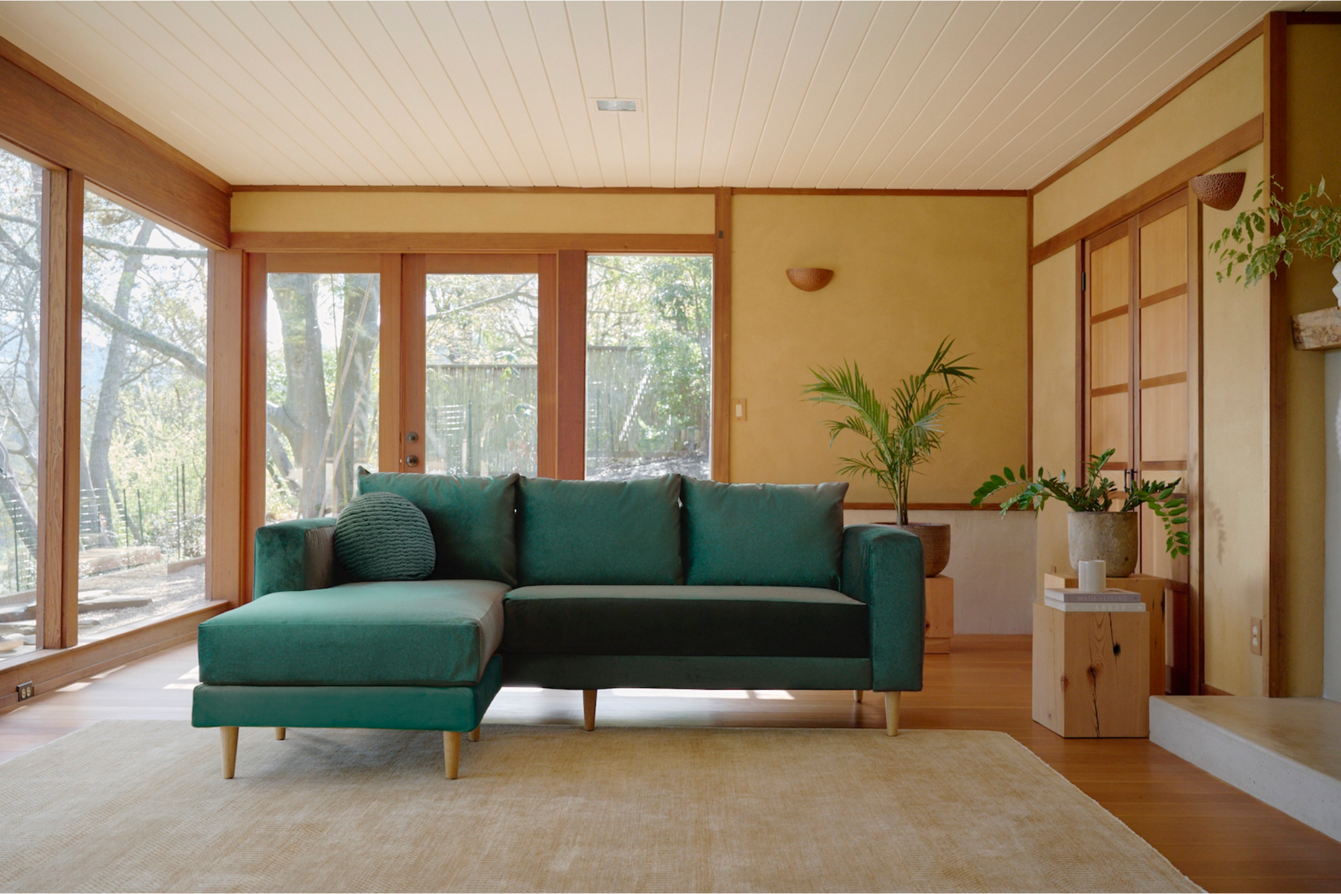 A modern living room showcases The Essential Sectional in a green fabric, elegantly positioned on a beige rug. Large windows with wooden frames provide a stunning view of the lush greenery outside. The space is enhanced with wooden accents, a small potted plant, and soft lighting, fostering a cozy and inviting atmosphere.