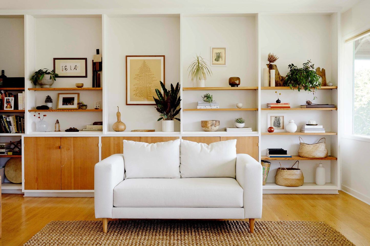 A cozy living room with The Essential Loveseat in white positioned in front of a wooden bookshelf adorned with various decorations, plants, books, and framed art. The room exudes a natural aesthetic featuring a jute rug and is illuminated by bright natural light streaming through a large window.