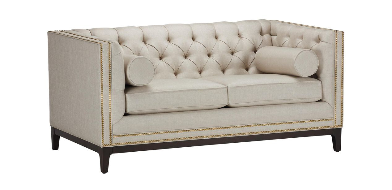 The Anderson Loveseat is a beige, tufted piece with a high back and sides, featuring two seat cushions adorned with decorative brass nailhead trim. It comes with two cylindrical bolster pillows and stands on dark wooden legs.