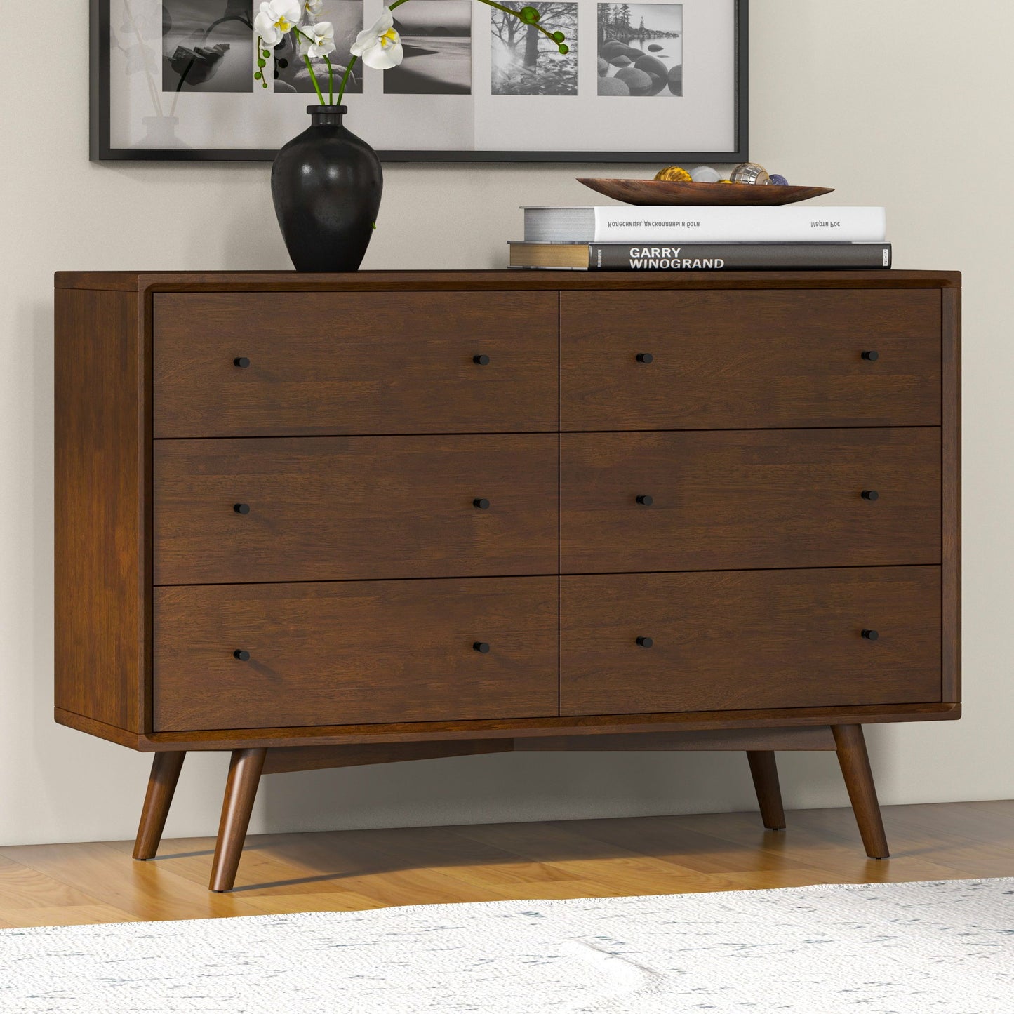 The Caroline mid-century modern solid wood dresser in brown features six drawers and tapered legs, topped with a black vase of white flowers and stacked books. Black-and-white photos adorn the wall above, while the dresser sits on light wooden flooring.