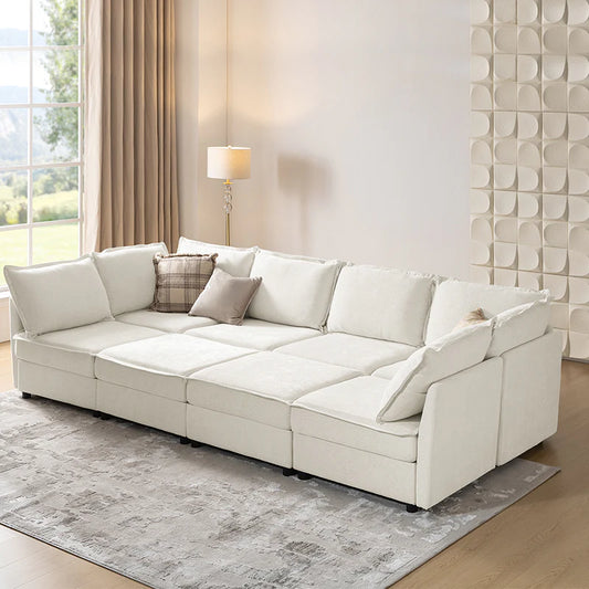 A modern living room showcases the Victoria Oversized Soft Sectional Sofa with Storage in a cream color, complemented by multiple cushions and a plaid throw pillow. A floor lamp stands nearby, while an intricate wall design adds visual interest. Sunlight filters through tall windows, illuminating the textured gray rug on the wooden floor.