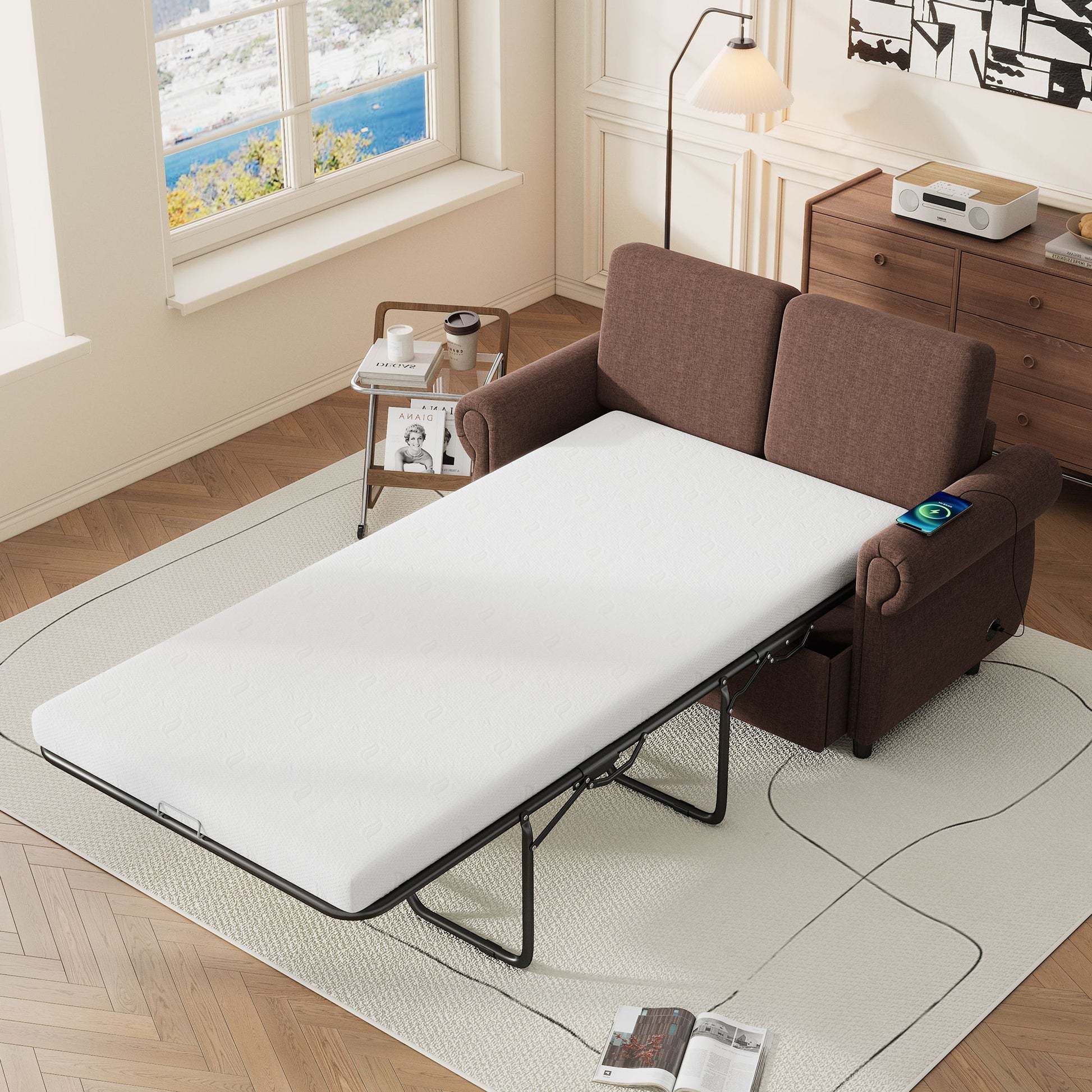 A Pull Out Sofa Bed Loveseat Sleeper with a twin-size memory mattress and two USB ports unfolds in a cozy living room with parquet floor. Its white mattress extends toward an ocean-view window, while a side table holds books and a lamp, and a wooden cabinet stores decor items and a radio.