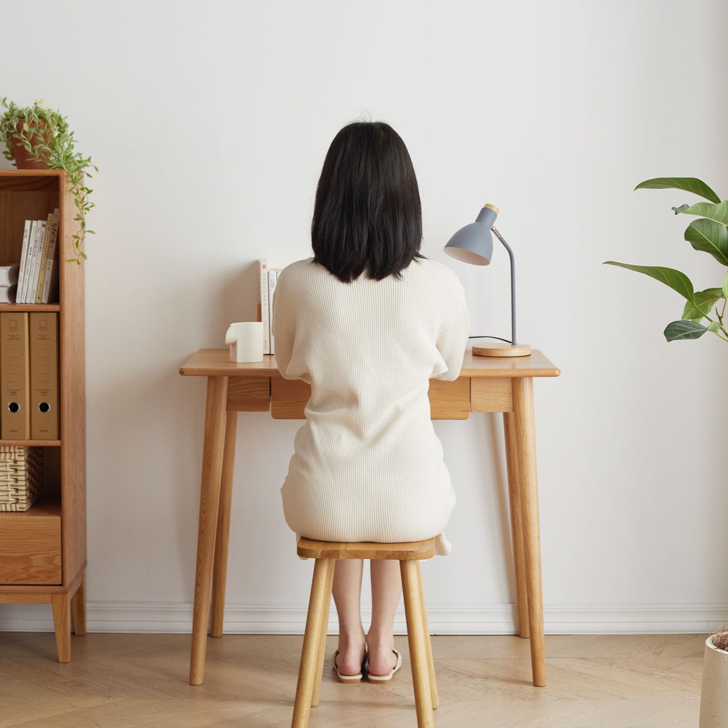 A woman with shoulder-length black hair sits on a wooden stool at an oak natural computer desk, featuring a drawer. The desk has a lamp and books, while green plants decorate the space and shelves hold files and baskets. The room is finished with wooden flooring and white walls.