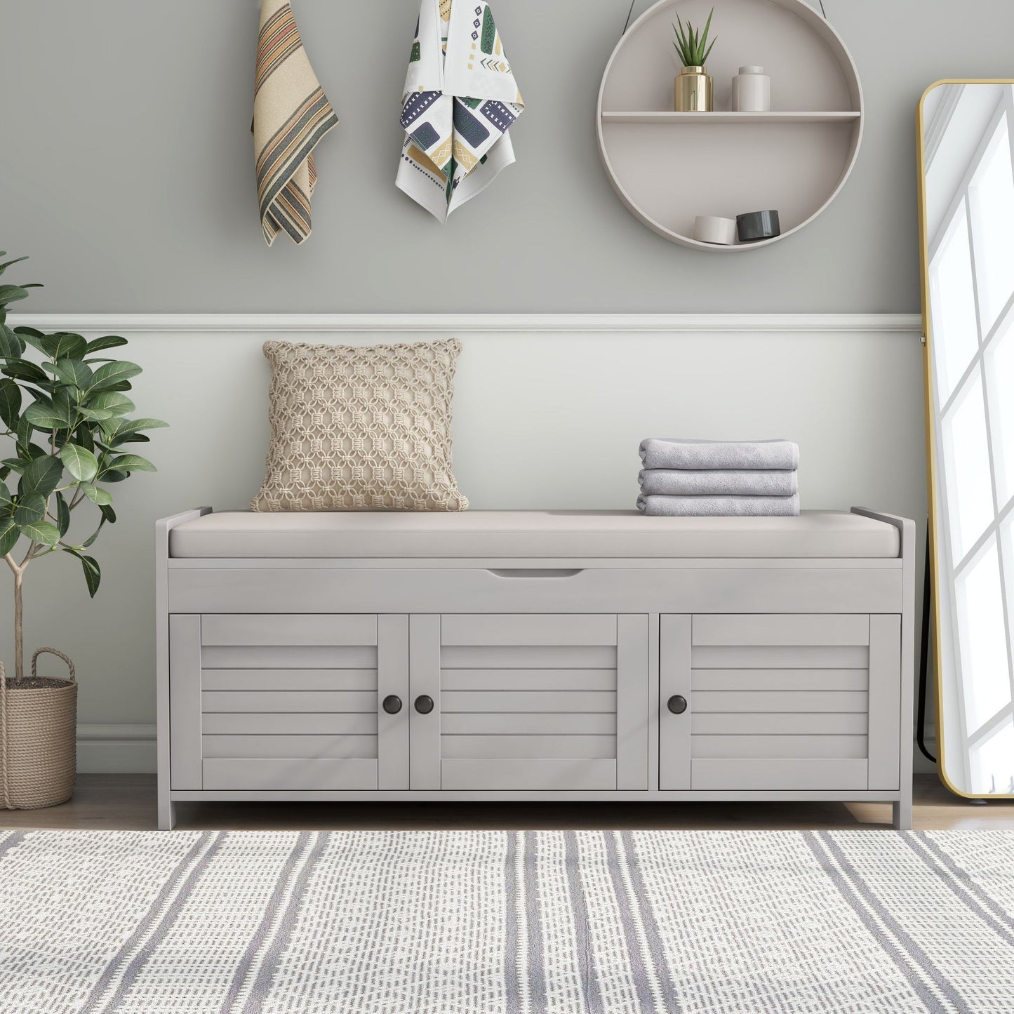 A minimalist room includes a light gray storage bench with shutters and a removable cushion, topped with folded towels. A textured pillow leans on the wall. A round shelf, tall mirror, hanging towels, a potted plant adorn the space. A striped rug covers the floor.