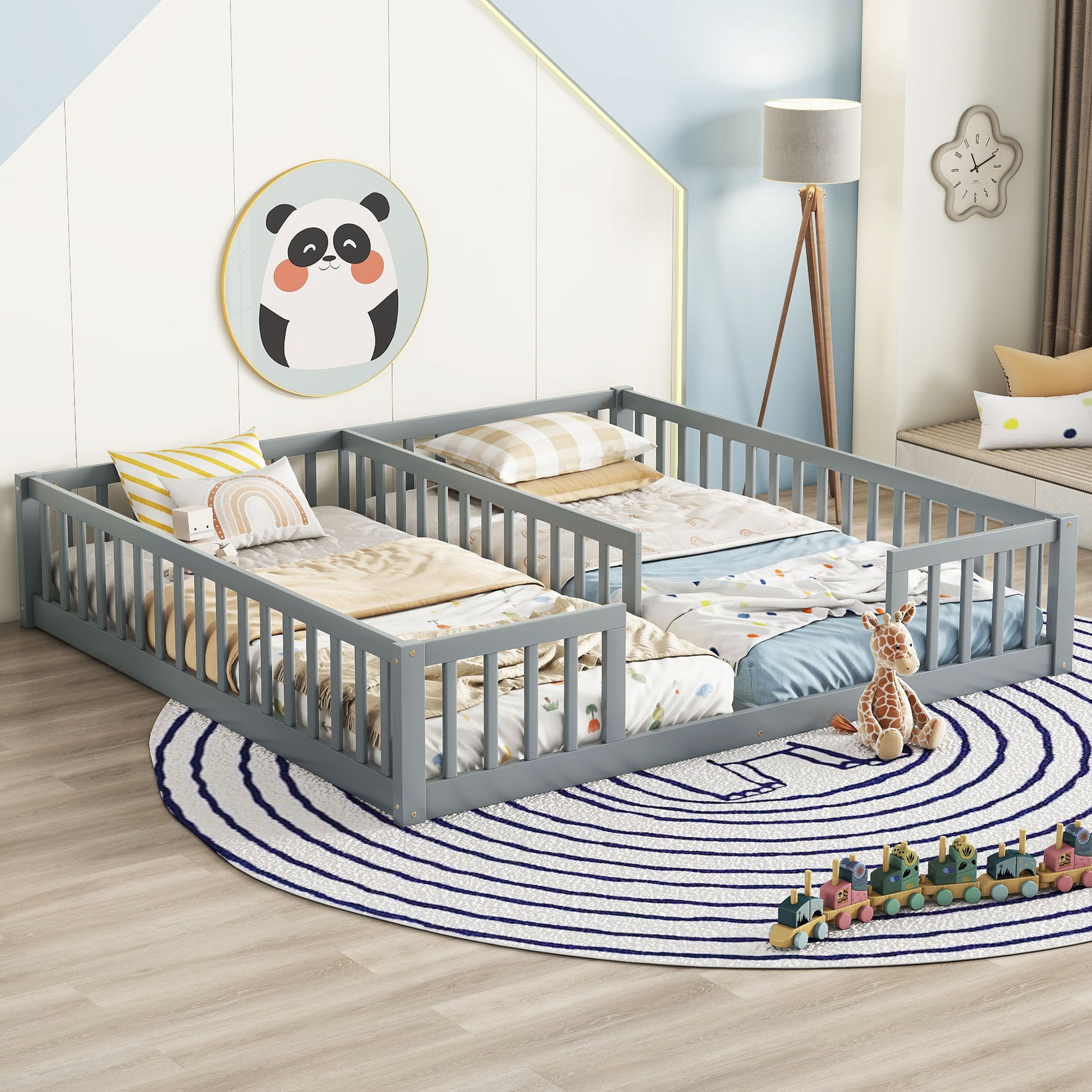 A children's bedroom features two gray Double Floor Beds With Fence and colorful bedding. A round spiral-patterned rug lies underneath, accompanied by a giraffe plush toy and small train set. Panda wall decoration and a lamp complete the cozy space.