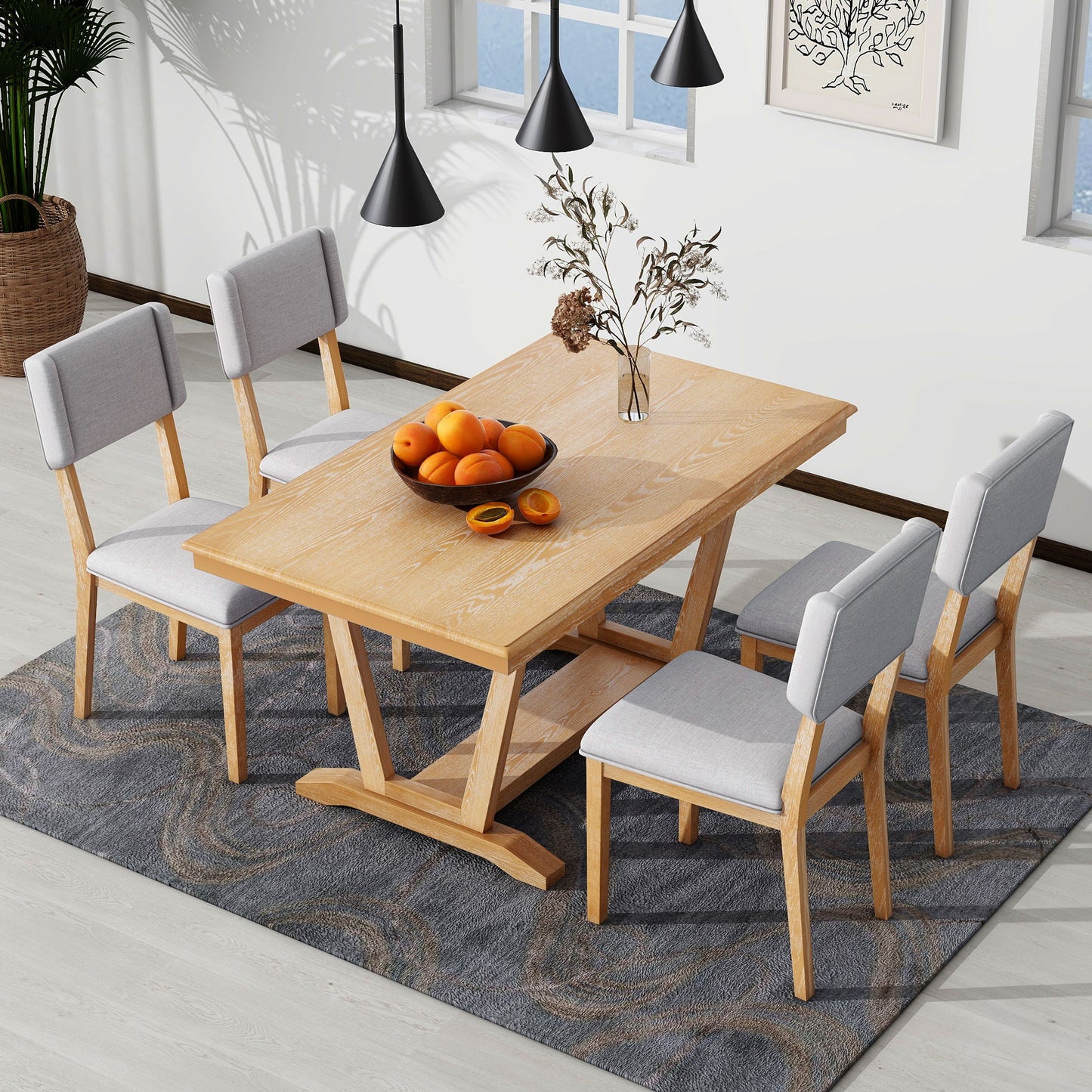 The Topmax 5-Piece Rustic Dining Table Set includes a rectangular dining table with a trestle base and four upholstered chairs, all on a gray patterned rug. A bowl of oranges and dried flowers sit atop, beneath three black pendant lights, beside framed art and two windows.