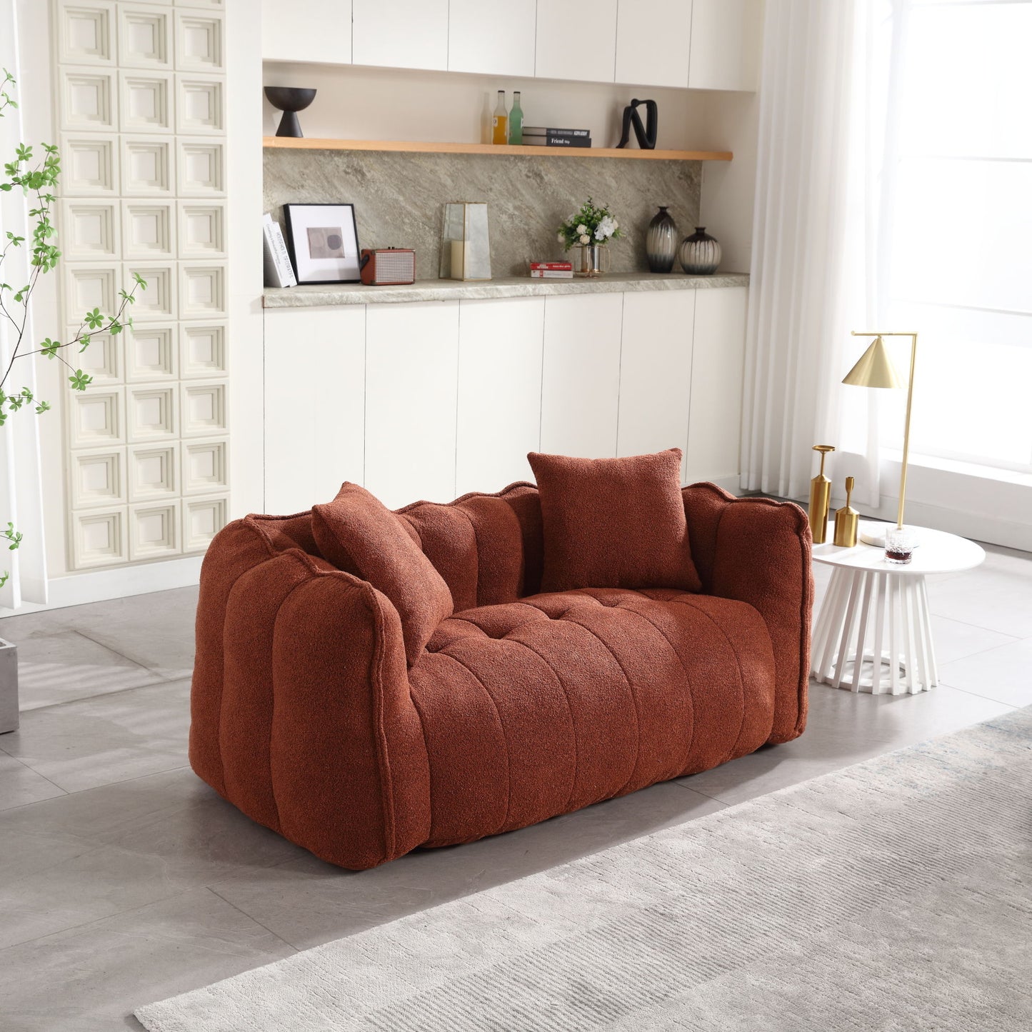 A cozy rust-colored tufted sofa with two cushions sits in a modern living room featuring a light-toned rug, white side table with a gold lamp, and decor-laden shelves against a textured wall. Sunlight fills the space through a window, complementing the soft beanbag chair for two with resilient foam.