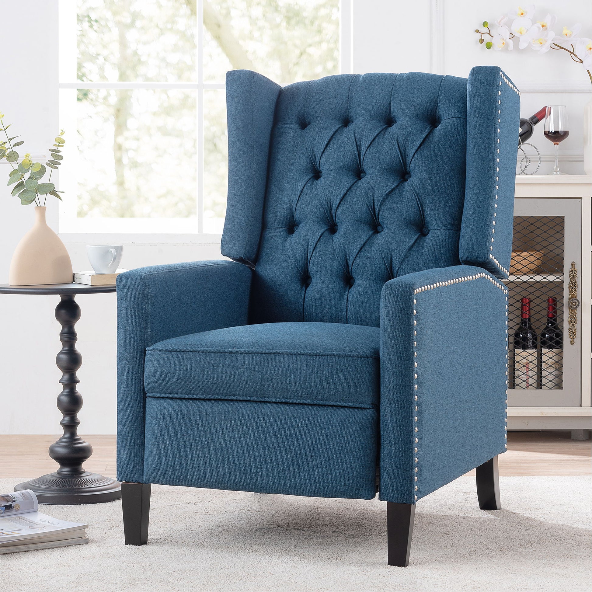 The Manual Wing Chair Recliner features a tufted backrest and silver nailhead trim. It's placed beside a small black side table adorned with a vase and plant, while natural light pours in through the window behind.