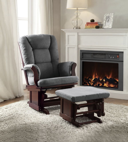 A cozy living room scene with the Adeliza Microfiber Glider Chair and Ottoman in gray on a fluffy beige rug, placed before a white fireplace with a flickering fire. A lamp and books adorn the mantel while natural light streams through sheer curtains.