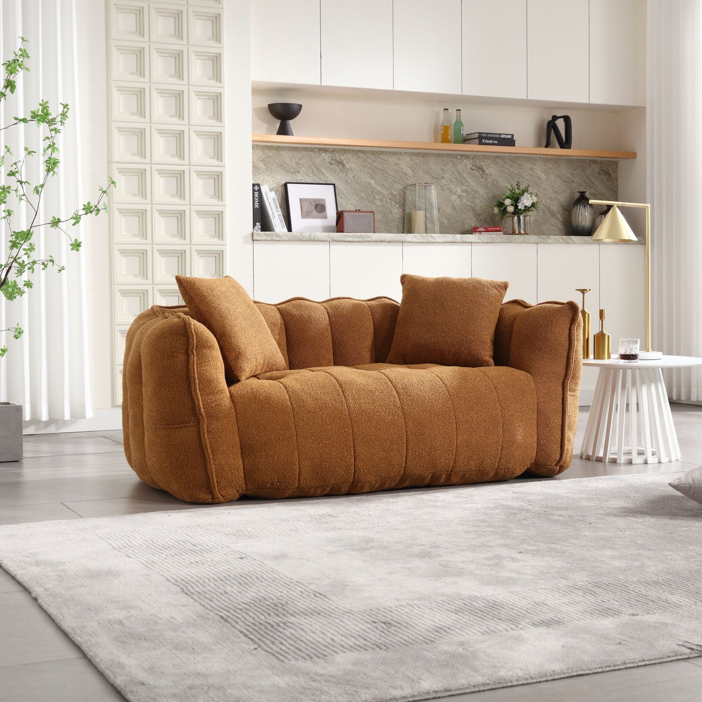 A modern living room showcases a plush brown sofa with two cushions on a light gray rug. Behind it, built-in white cabinets and shelves hold decorative items. To the right, there's a small white table.