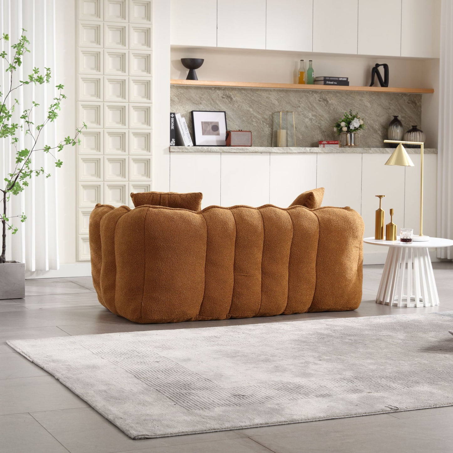 A cozy, brown tufted sofa sits in a modern living room with light gray flooring. A textured gray rug and round white coffee table with a gold lamp enhance the decor, alongside a Soft Beanbag Chair With High Resilience Foam Core For Two People. White cabinetry and a stone backsplash complete the scene.