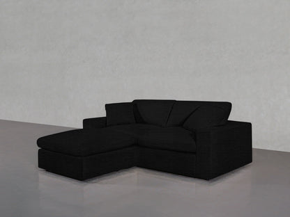 A modern 3-Seat Modular Chaise Sectional in a dark charcoal shade is positioned against a plain light gray wall on a polished concrete floor, featuring two back cushions and an extended chaise for lounging.