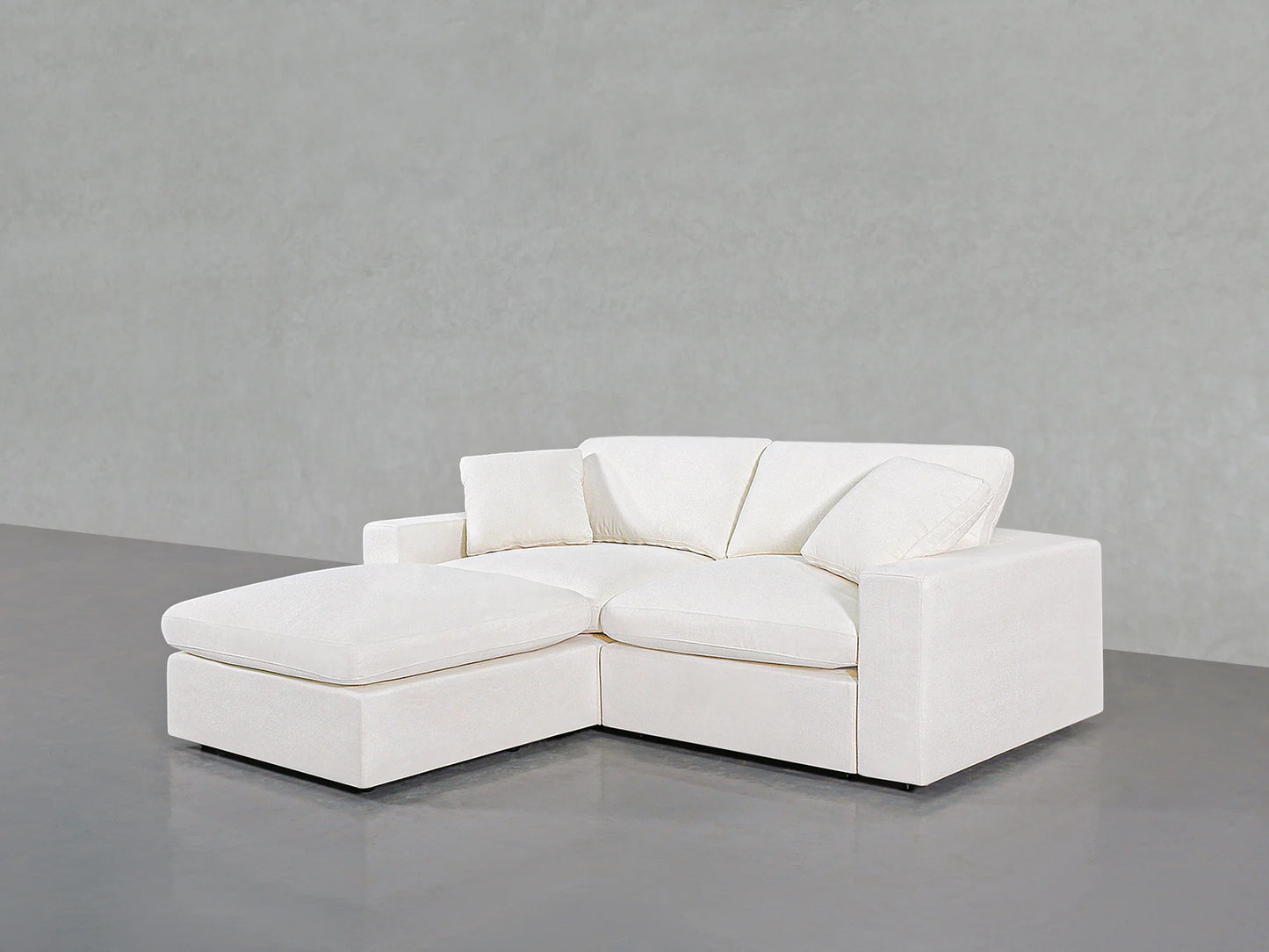 A contemporary 3-Seat Modular Chaise Sectional in white, featuring clean lines and plush cushions, is perfectly positioned on a sleek grey floor against a plain grey wall. This sectional showcases an L-shaped design with a chaise lounge on the left side.