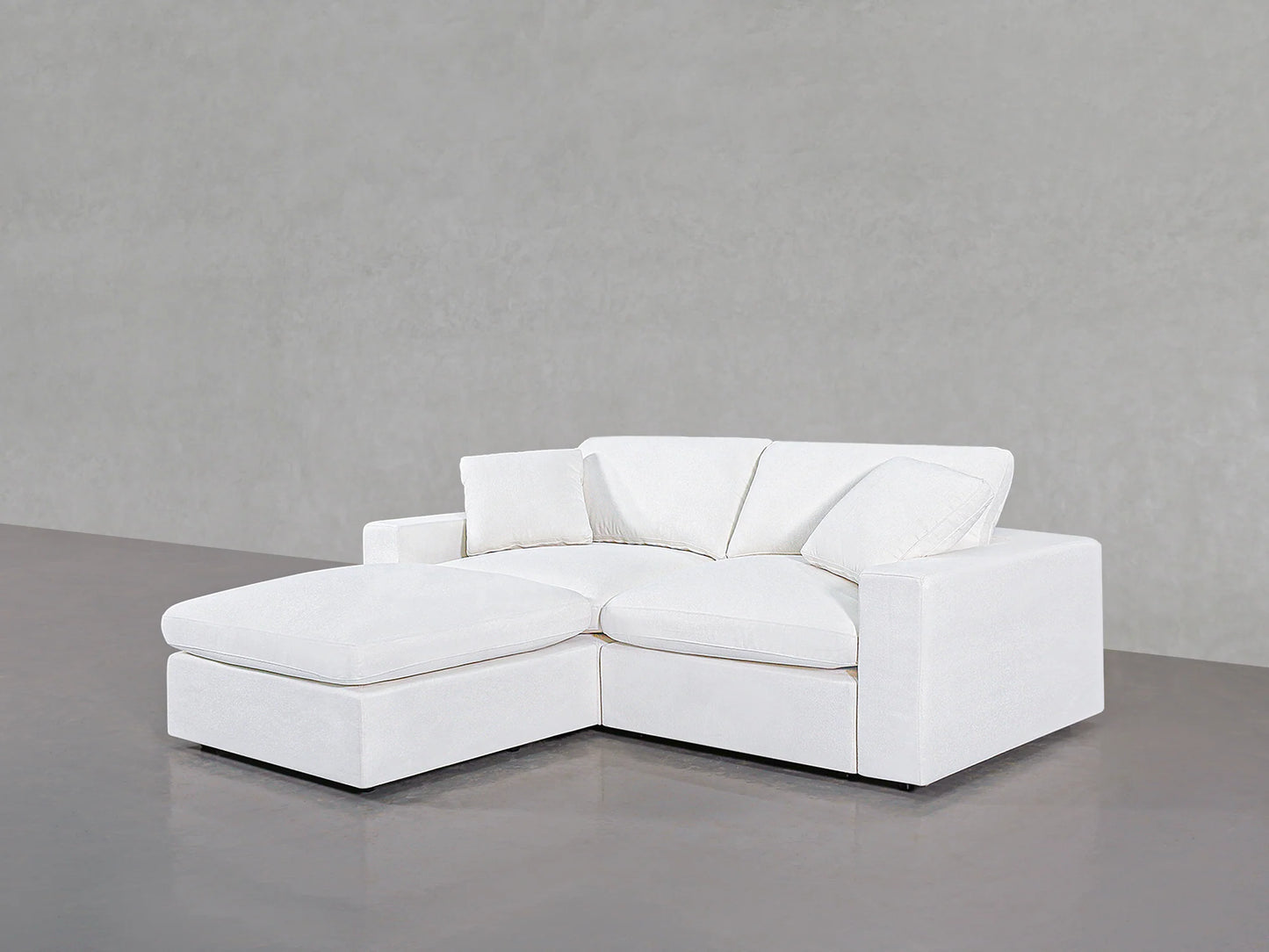 A 3-Seat Modular Chaise Sectional in a minimalist white design, featuring a chaise lounge on the left side, is set against a plain gray background. This sofa boasts clean lines and includes three matching throw pillows.