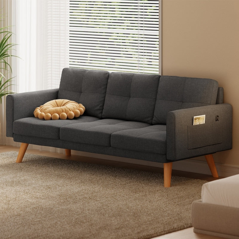 A modern living room showcases a dark gray Sofa Couch Small Loveseat, highlighted by its wooden legs and featuring three comfy pillows. A round, croissant-shaped pillow is gracefully positioned on the loveseat, which also includes a convenient arm pocket. Complementing the scene is a beige carpet and a nearby plant beside the window adorned with blinds.