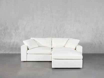 A 3-Seat Modular Chaise Sectional in a sleek white design, complete with plush cushions, is set against a textured gray wall on a polished concrete floor. The sectional includes a left-arm loveseat paired with an adjoining chaise lounge, accentuating the minimalist decor that highlights this striking piece of furniture.