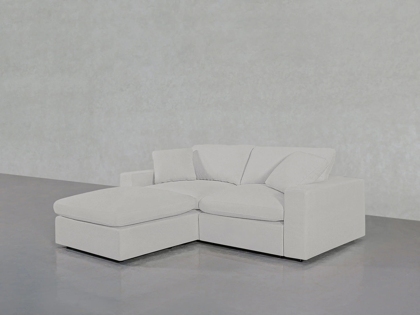 A contemporary 3-seat Modular Chaise Sectional, with an L-shaped configuration and the chaise lounge on the left side, is positioned on a sleek, reflective gray floor against an understated gray backdrop. The sofa boasts clean lines and a minimalist design, featuring plush cushions.