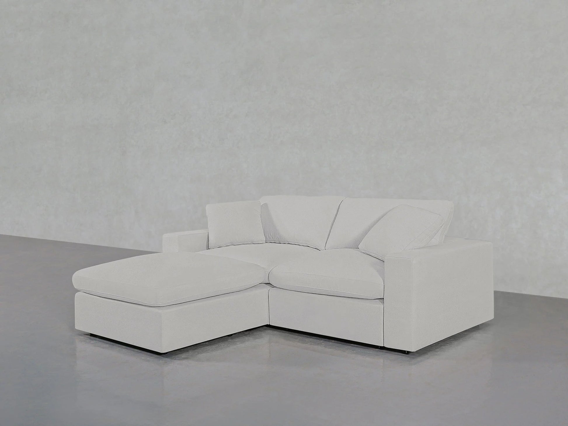 A contemporary 3-seat Modular Chaise Sectional, with an L-shaped configuration and the chaise lounge on the left side, is positioned on a sleek, reflective gray floor against an understated gray backdrop. The sofa boasts clean lines and a minimalist design, featuring plush cushions.