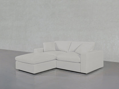 A contemporary 3-seat Modular Chaise Sectional, with an L-shaped configuration and the chaise lounge on the left side, is positioned on a sleek, reflective gray floor against an understated gray backdrop. The sofa boasts clean lines and a minimalist design, featuring plush cushions.