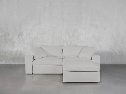 The 3-Seat Modular Chaise Sectional, featuring a modern design and light gray upholstery, is positioned against a neutral textured wall on a smooth concrete floor. It is equipped with plush cushions and includes a chaise lounge extension on the right side.