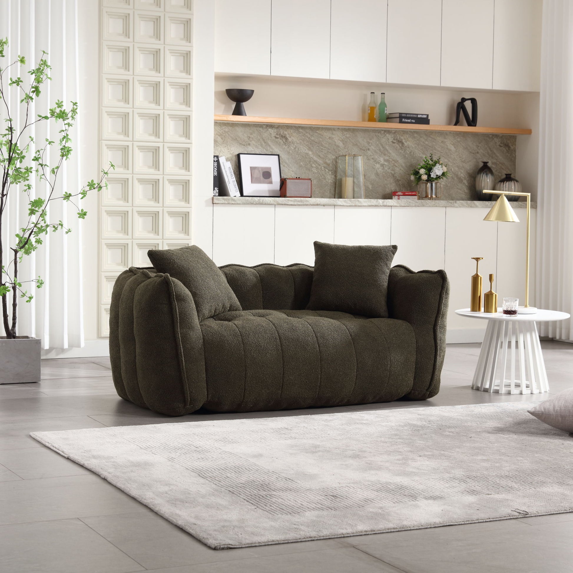 A modern living room includes a dark green, plush sofa with two cushions on a light gray rug. A white cabinet, decorative plant, and small round white table with a golden lamp and books complement the setting. For additional seating, there's a soft beanbag chair for two with a high-resilience foam core.