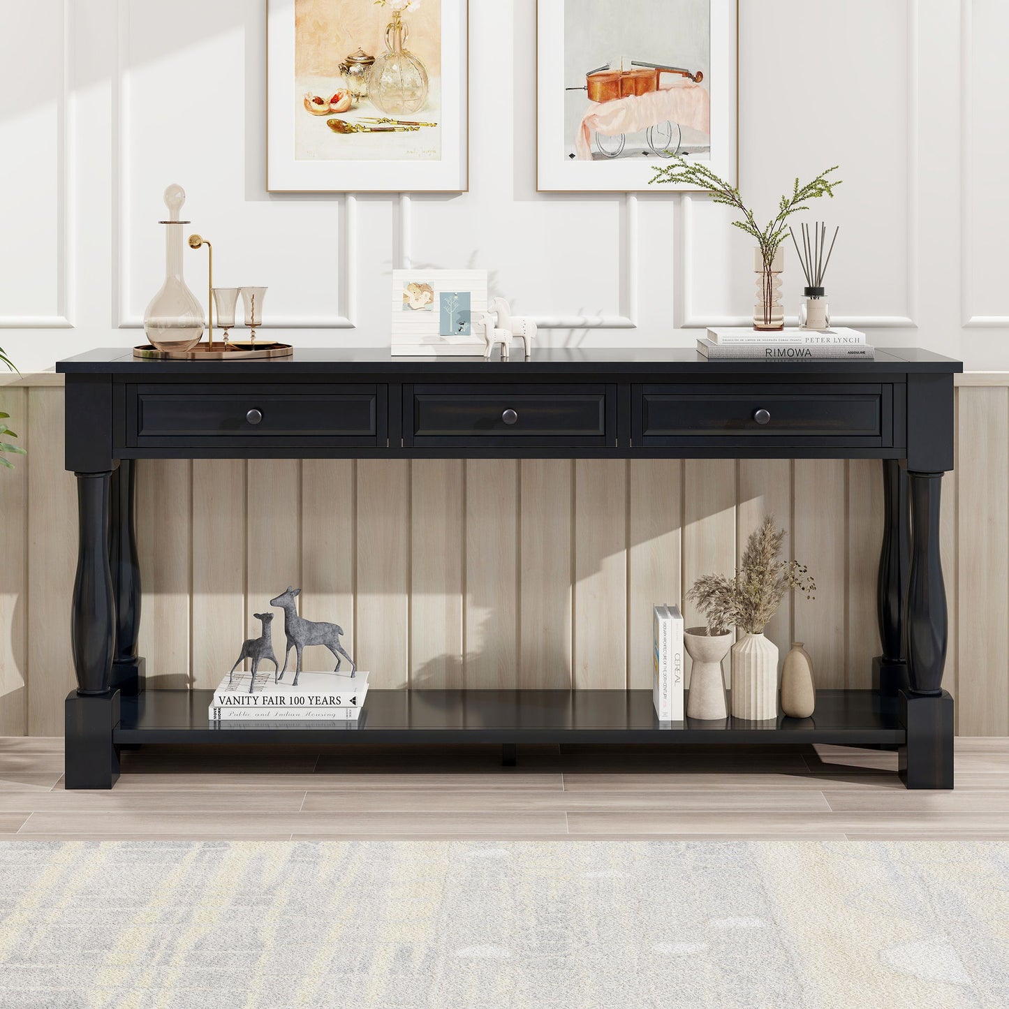 A black console table with three drawers and a shelf, ideal for an entryway, hallway, or living room, stands against a light-paneled wall. It's adorned with books, flowers in a vase, a horse figurine, and framed art above. The room features a light rug and wooden floor.