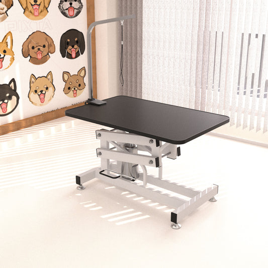 The Hydraulic Dog Pet Grooming Table, featuring a black surface and adjustable metal frame, sits in a sunlit room. A curtain and a poster with cartoon dog faces, including Shiba Inu illustrations, are displayed on the wall behind it.