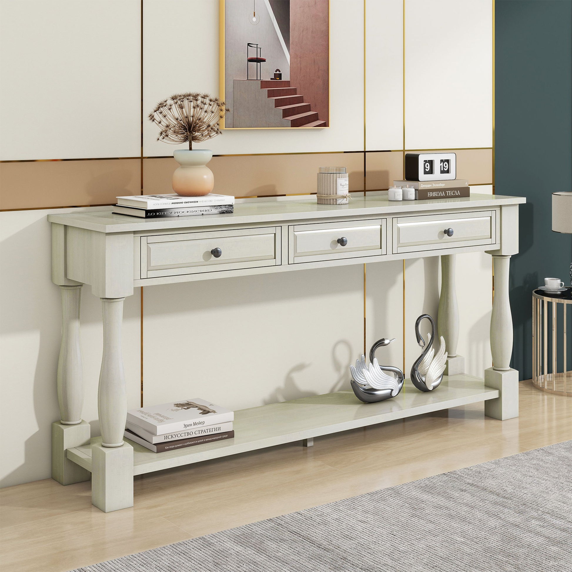 A cream-colored long console table with three drawers is adorned with books, a vase with dried flowers, and two silver swan figurines. It stands against a modern geometric-patterned wall featuring small framed artwork.