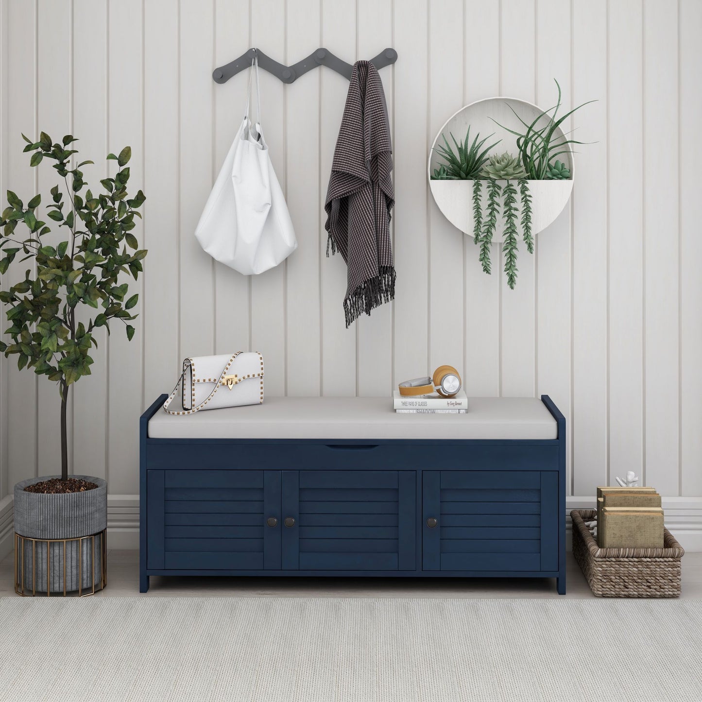 A stylish entryway features a blue storage bench with 3 shutter-shaped doors, a removable cushion, and hidden storage. A potted tree and wicker basket are nearby. Above, a coat rack holds a white bag and checkered scarf. A circular wall planter with greenery decorates the light gray wall.