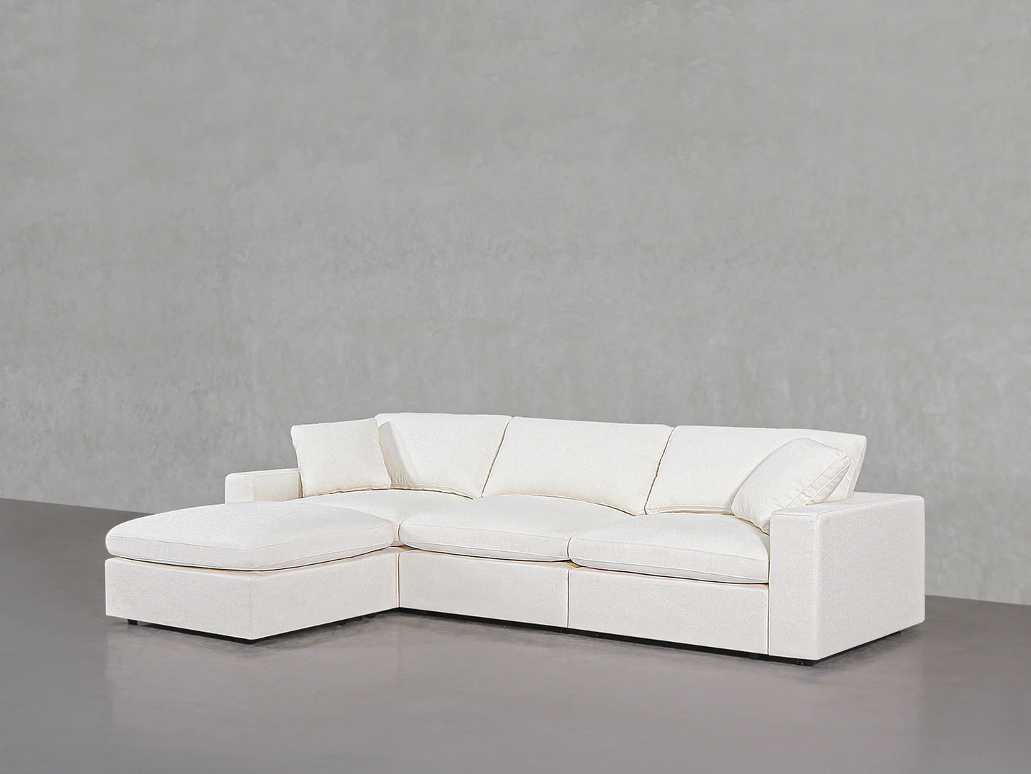 A 4-Seat Modular Chaise Sectional in sleek white, featuring clean lines and plush cushions, is displayed against a simple gray background. This sofa includes a left-side chaise, providing a roomy and comfortable seating area.