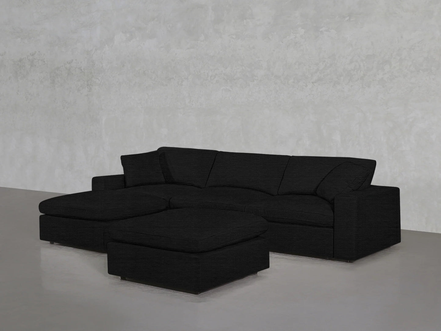 A 4-Seat Modular Chaise Sectional with Ottoman, featuring minimalist and modern design, is placed against a textured gray wall. The clean lines and black upholstery contribute to a sleek and contemporary appearance on the polished concrete floor.