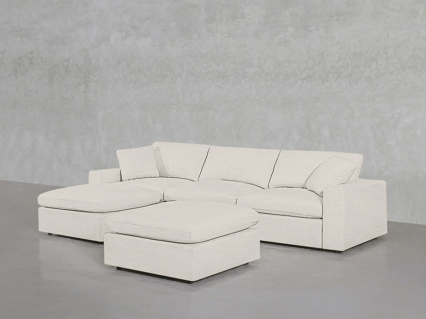 A 4-Seat Modular Chaise Sectional with Ottoman in a neutral hue is placed against a textured gray wall. The furniture features clean lines and a modern design, sitting on a plain gray floor.